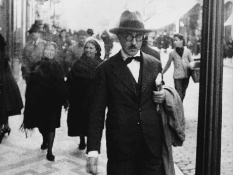 Fernando Pessoa walks on a busy sidewalk in the Baixa district of Lisbon, Portugal. He wears a fedora hat, a bow tie under a white collar, and a long black trench coat. He carries a book.