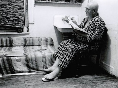 black and white photo of Philip Whalen sitting in a chair next to a sink and couch, reaching a book.